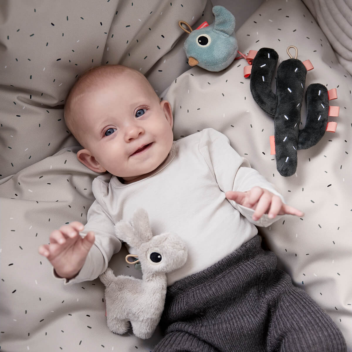 Baby lays with Done by Deer soft toys Lalee and Birdee on bedlinen in sand